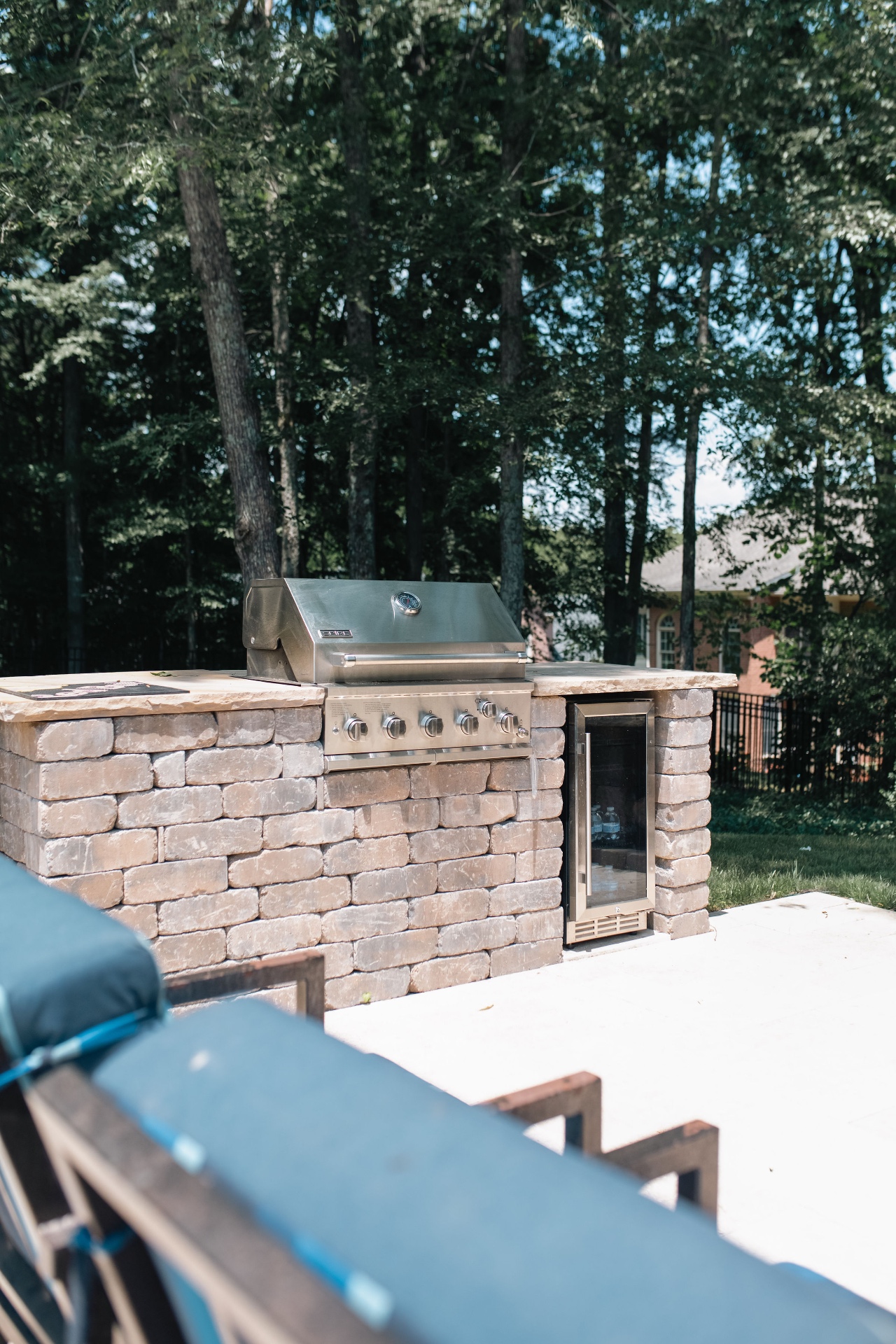 Stone outdoor kitchen with stainless gas grill and built-in beverage refrigerator on the poolside patio.