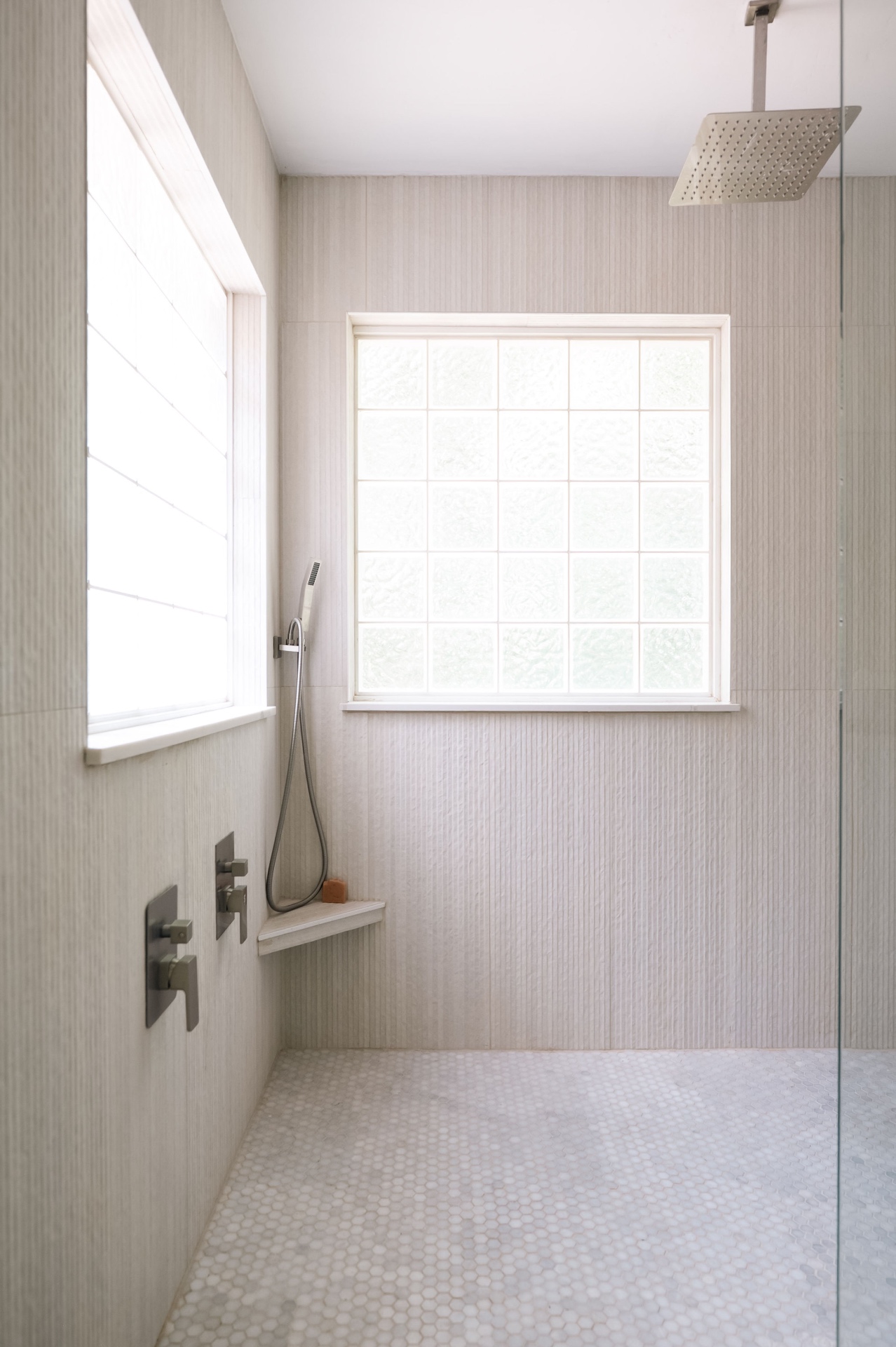 Oversized walk-in primary shower with rain head, hand shower, bench, and glass block window.