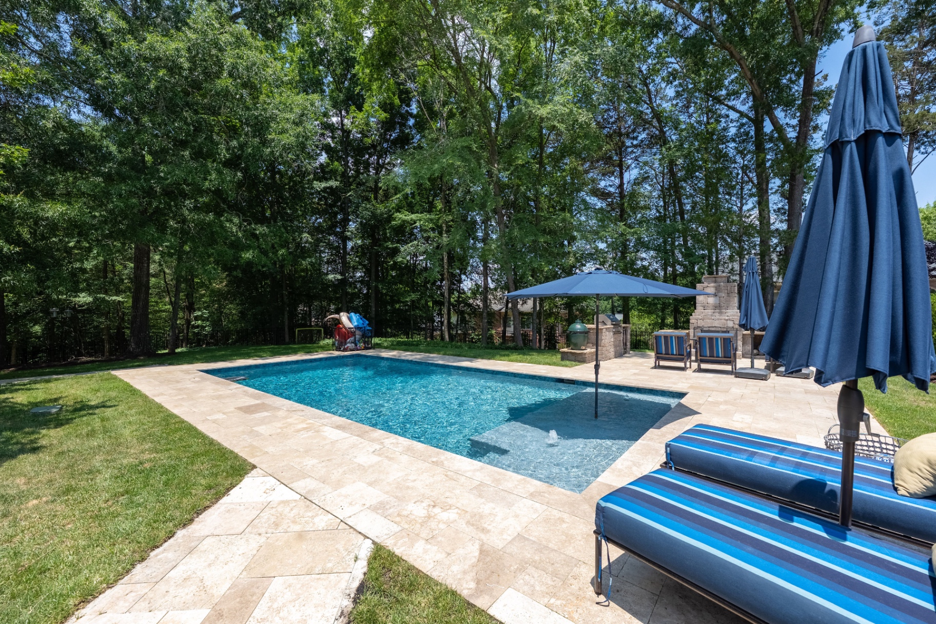 Saltwater pool with tanning ledge, expansive stone decking, and private wooded backdrop.